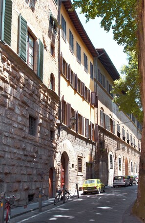 Un edificio de varios pisos con fachadas de piedra y enlucido, múltiples ventanas con contraventanas, y una calle con coches estacionados y una bicicleta.