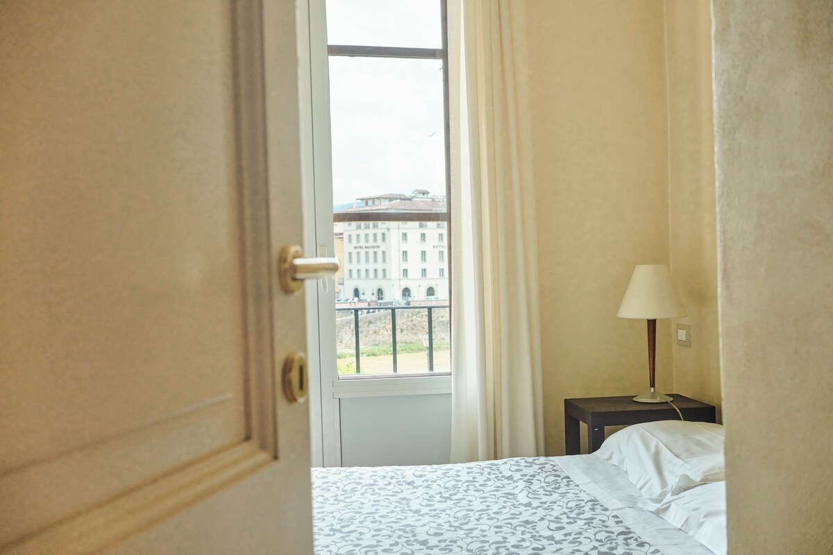A hotel room with a bed, a nightstand, a lamp, and a window with a view of a building.