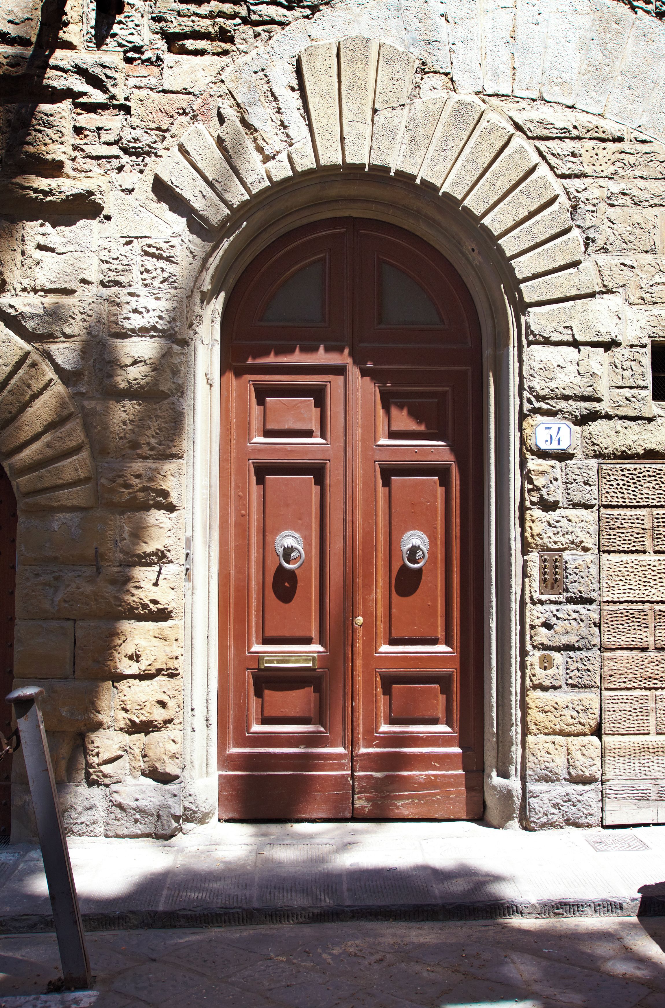 Una puerta doble de madera con un arco de piedra, un aldabón de metal y una placa con un número.