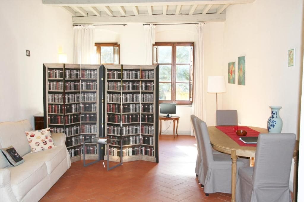 Uma sala de estar aconchegante com um sofá, uma mesa de jantar com cadeiras, uma estante de livros e uma janela com cortinas.
