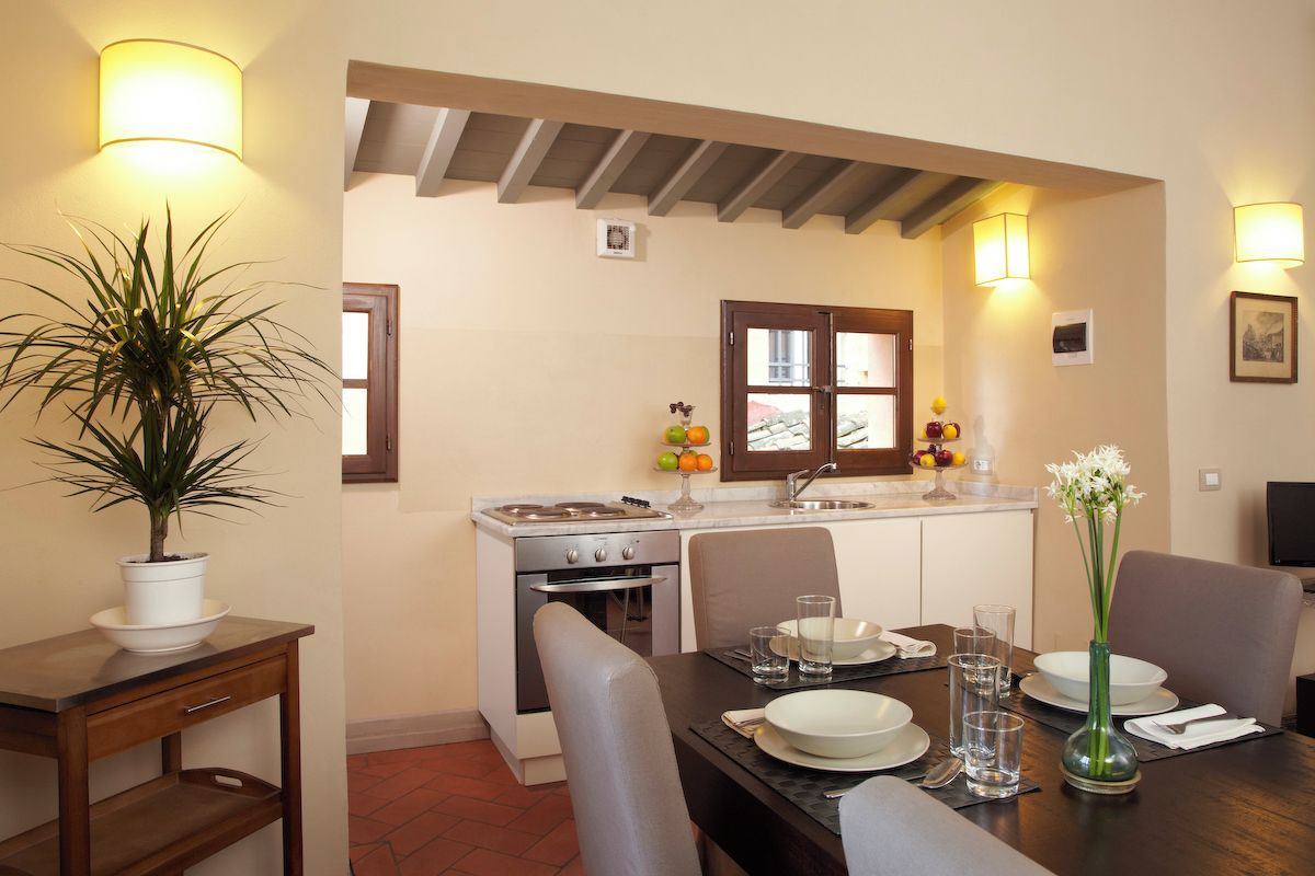 Un comedor con una mesa preparada para dos, una planta en maceta y una cocina al fondo.
