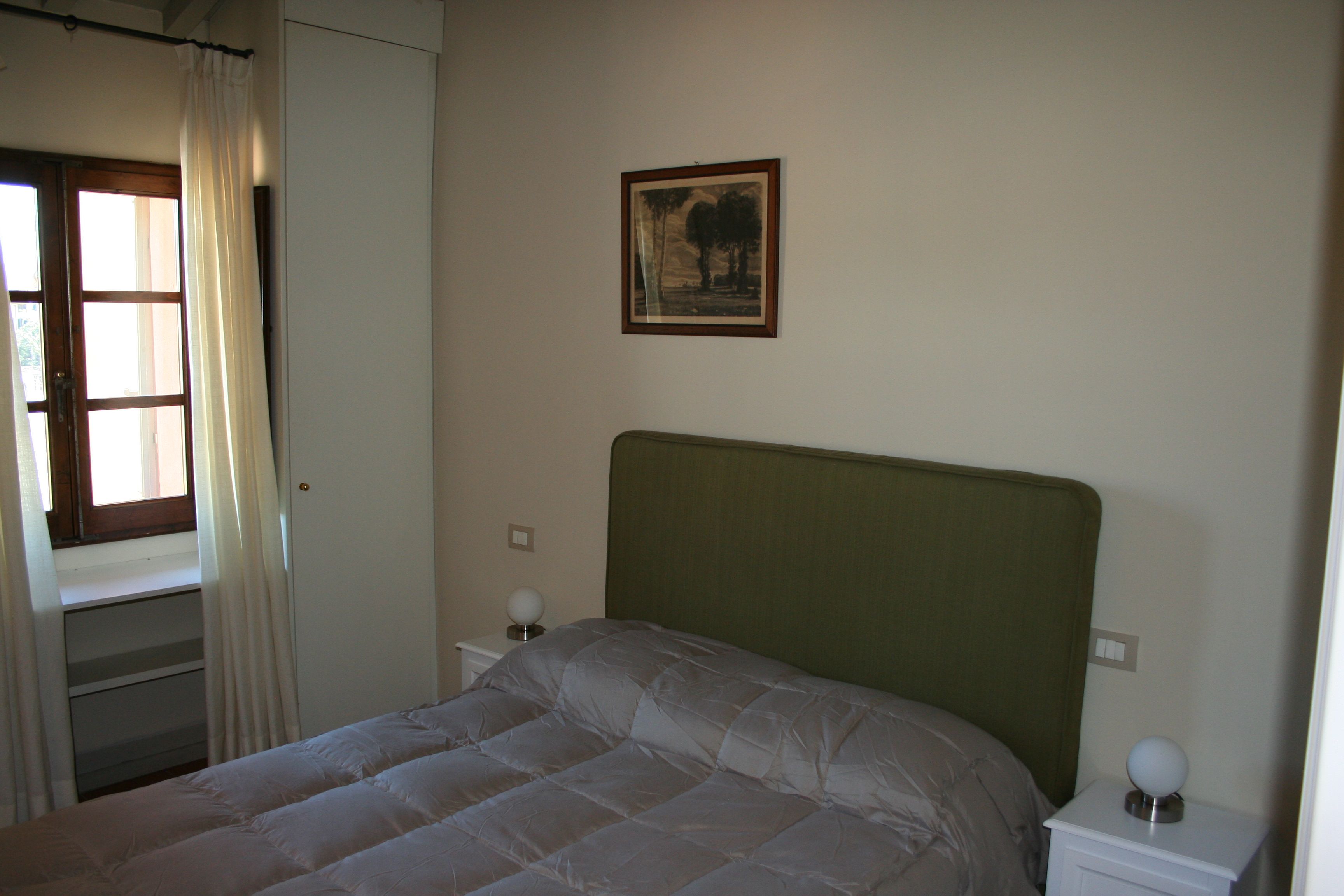 A bedroom with a bed, a nightstand, a wall-mounted lamp, a window with curtains, and a framed picture on the wall.