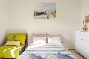 Une chambre à coucher comprenant un lit, une table de chevet, une commode et un tableau représentant une plage, accroché au mur.