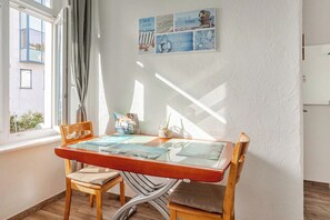 Un coin repas avec une table et des chaises en bois, une fenêtre avec des rideaux et un mur orné d’une affiche sur le thème de la plage.