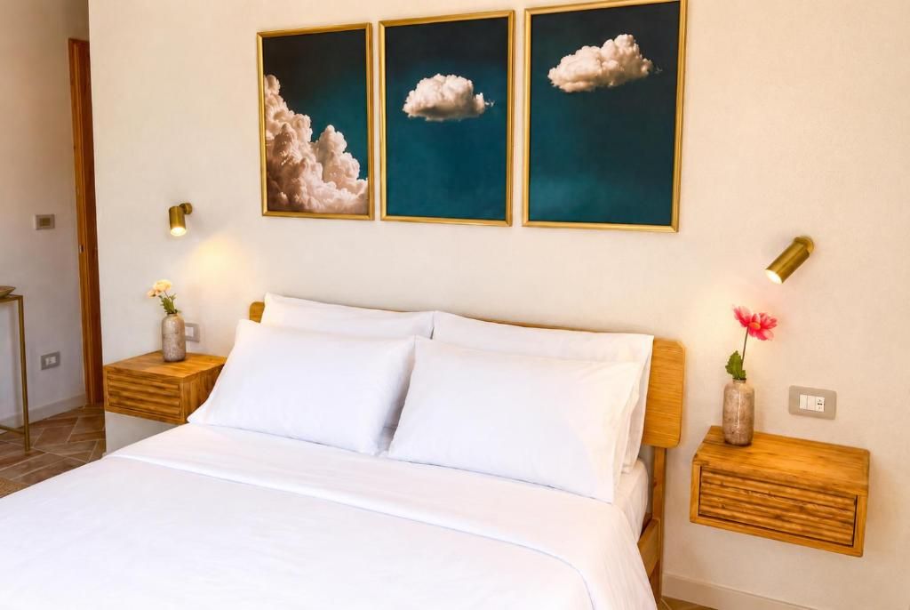Ein Hotelzimmer mit einem Bett, zwei Nachttischen, einer Blumenvase und drei gerahmten Gemälden mit Wolkenmotiven an der Wand.