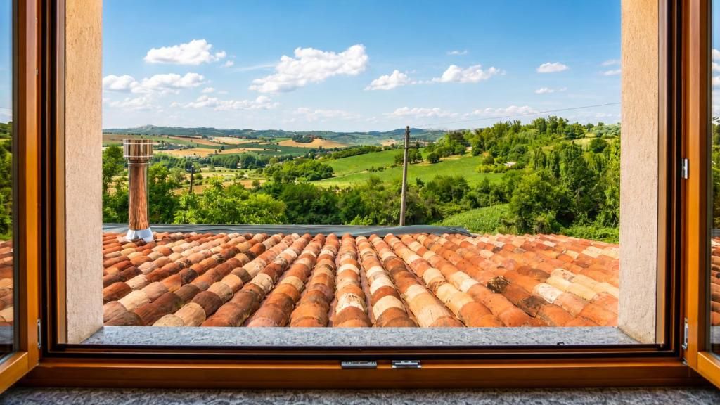 Vista a través de una ventana con tejado de baldosas y un paisaje de campos verdes y colinas.