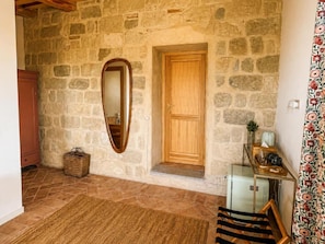 Interior de paredes de piedra con una puerta de madera, un espejo, una cesta de mimbre y una pequeña mesa con un vaso.