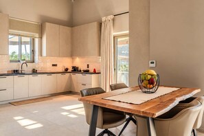 A modern kitchen with a dining area, featuring a wooden table and chairs, a fruit basket, and a view of the outdoors. - Villa Lydia, Luxury Stone Retreat with Private Pool (Agios Nikolaos)