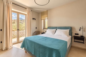 A bedroom with a bed, a nightstand, a lamp, and a view of the outdoors. - Villa Lydia, Luxury Stone Retreat with Private Pool (Agios Nikolaos)