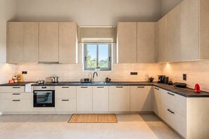 A modern kitchen with light-colored cabinets, a built-in oven, a sink, and a window with blinds. - Villa Lydia, Luxury Stone Retreat with Private Pool (Agios Nikolaos)