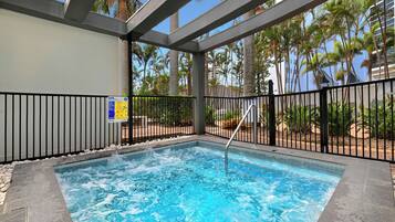 Un jacuzzi con barandilla metálica, rodeado de una zona de piscina con palmeras y un edificio al fondo.