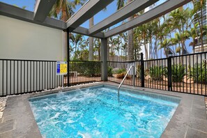 Un jacuzzi con barandilla metálica, rodeado de una zona de piscina con palmeras y un edificio al fondo.