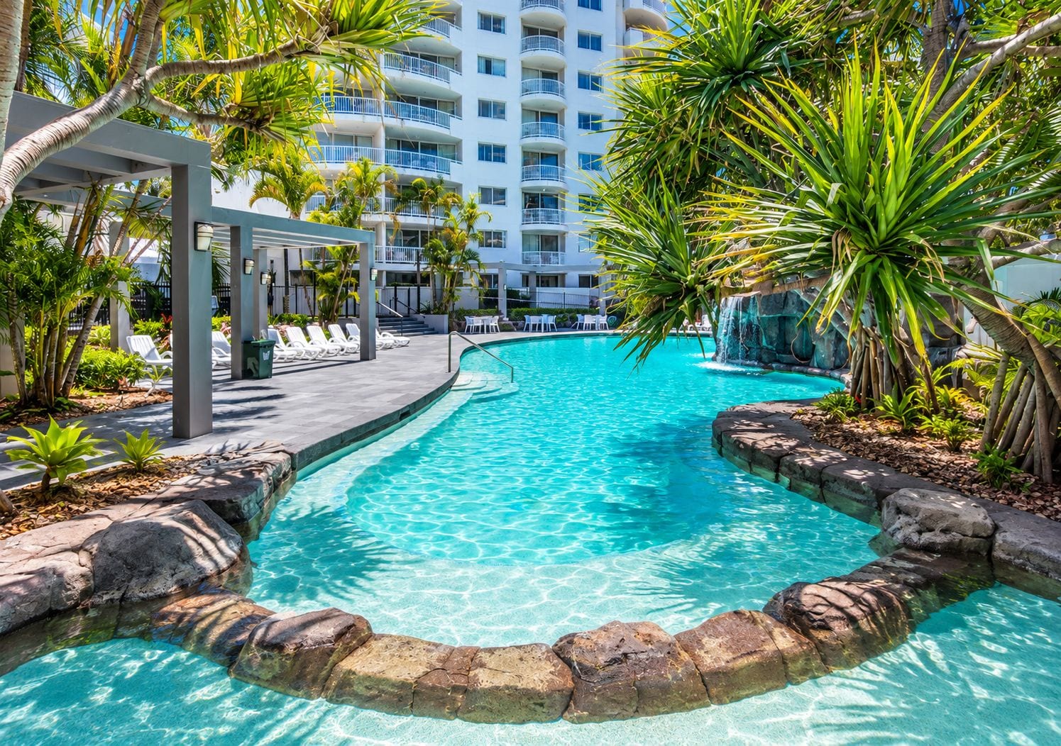 A clear blue pool with a stone border, surrounded by lounge chairs and palm trees, with a multi-story building in the background.