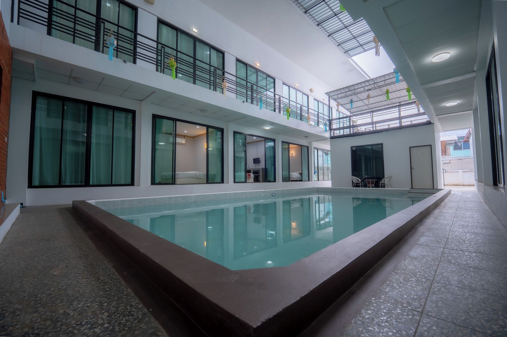 A modern indoor swimming pool with a glass barrier, multiple rooms with beds, and a balcony with railings.