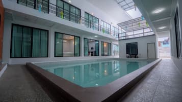 A modern indoor swimming pool with a glass barrier, multiple rooms with beds, and a balcony with railings.