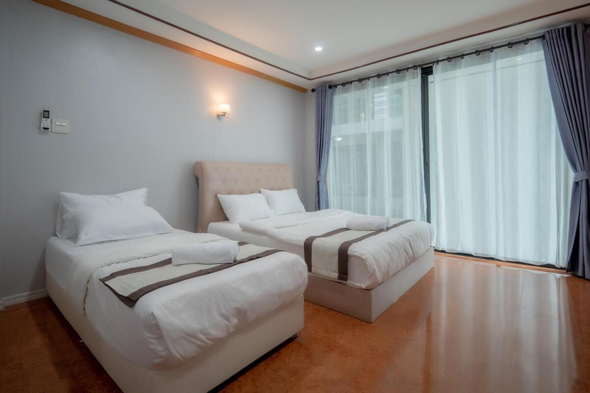 Habitación de hotel con dos camas, cabecero en tonos claros, un ventanal grande con cortinas y suelo de madera.