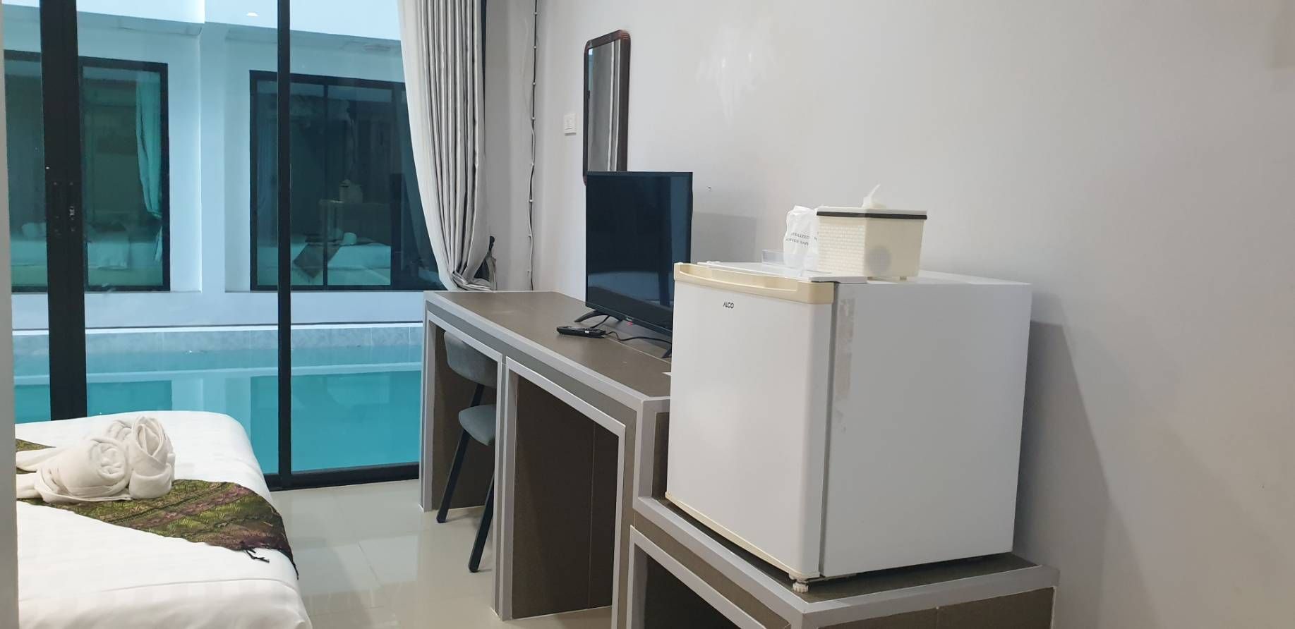 A hotel room with a bed, a desk with a computer, a refrigerator, and a window with a view of a pool.