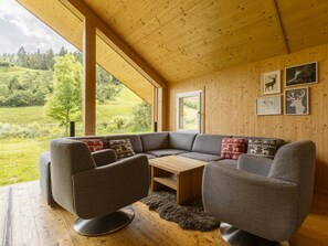 Un intérieur moderne avec un plafond en bois, un canapé d’angle gris, une petite table en bois et une vue sur un paysage verdoyant grâce à de grandes fenêtres.