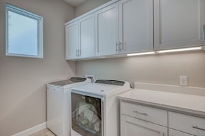 A laundry room with two front-loading washers and dryers, white cabinets above, and a window on the left. - The Bud Lane Collection | Infinity Pool, Dock & Waterslide (Kingsland)