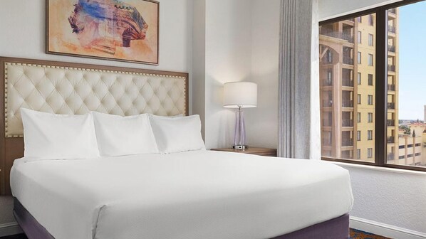 Quarto de hotel com uma cama grande, cabeceira, mesa de cabeceira com abajur, janela com vista para edifícios e um quadro na parede.