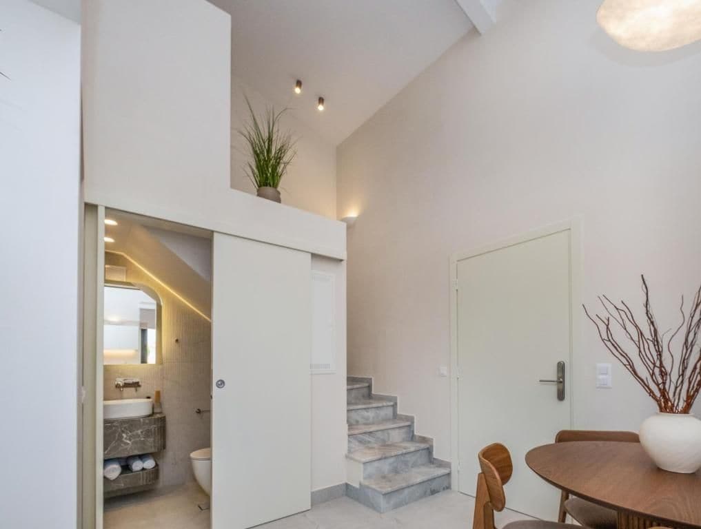 Un interior moderno con una escalera que lleva a un segundo piso, un comedor con mesa y sillas, y una puerta blanca.