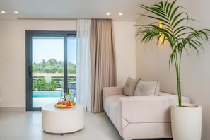 A modern living room with a sofa, a round coffee table with a fruit platter, a potted plant, and a view of a swimming pool through a glass door. - Calista Horizon by Booking Kottas (Kallithea)