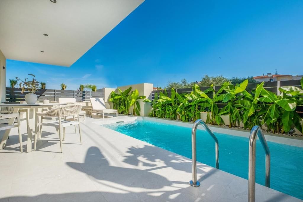 Un área de piscina en la azotea con un cielo azul despejado, rodeada de exuberante vegetación y mobiliario exterior blanco.