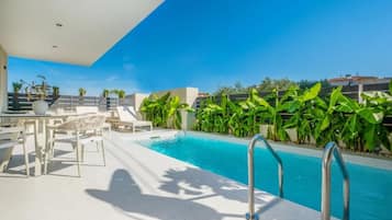 Un área de piscina en la azotea con un cielo azul despejado, rodeada de exuberante vegetación y mobiliario exterior blanco.