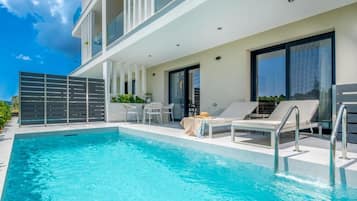 Una piscina moderna al aire libre con sillones de descanso y una mesa, rodeada por un edificio con balcones y ventanas grandes.