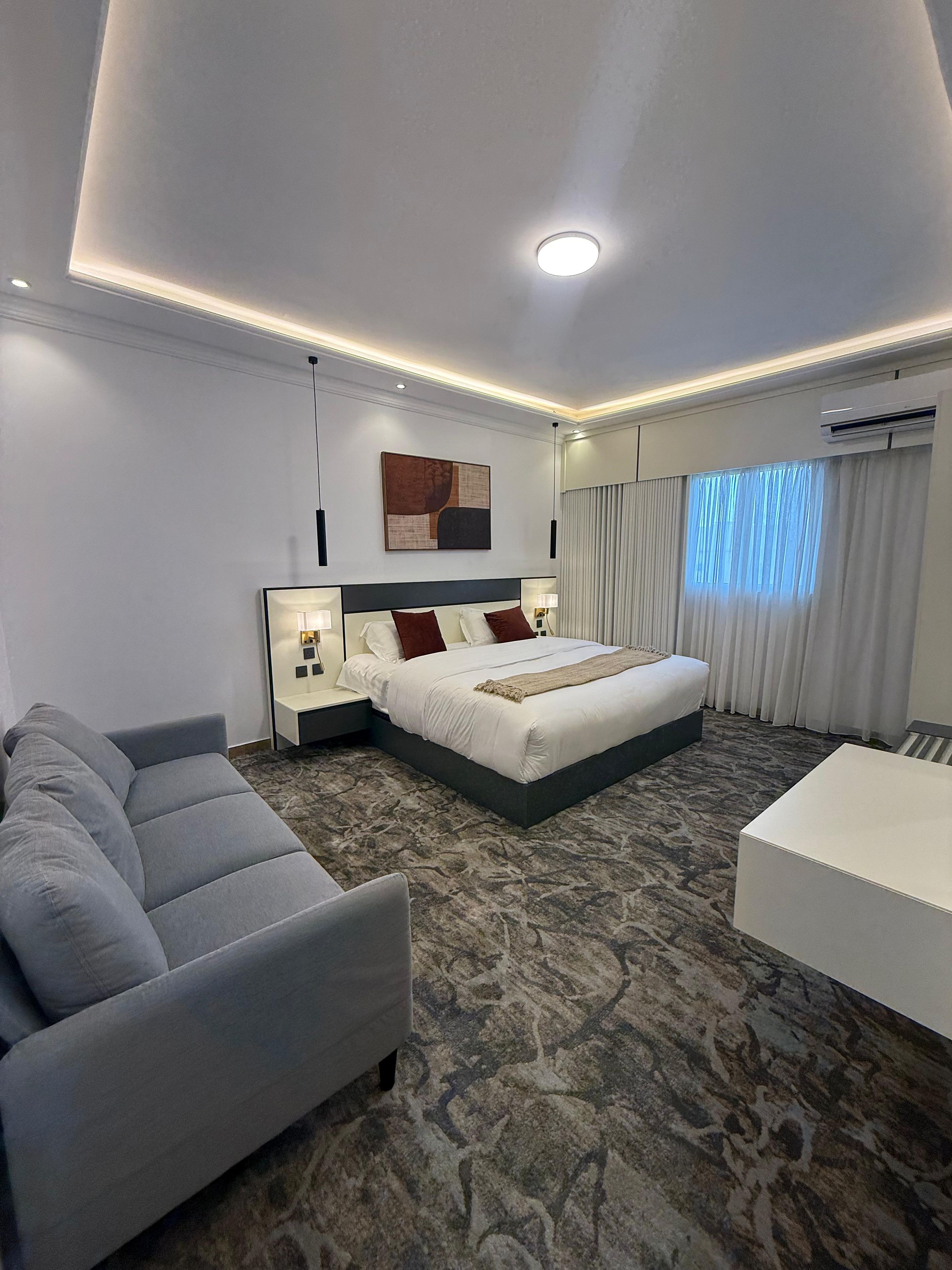 A modern hotel room with a large bed, a sofa, a small table, and a ceiling light.