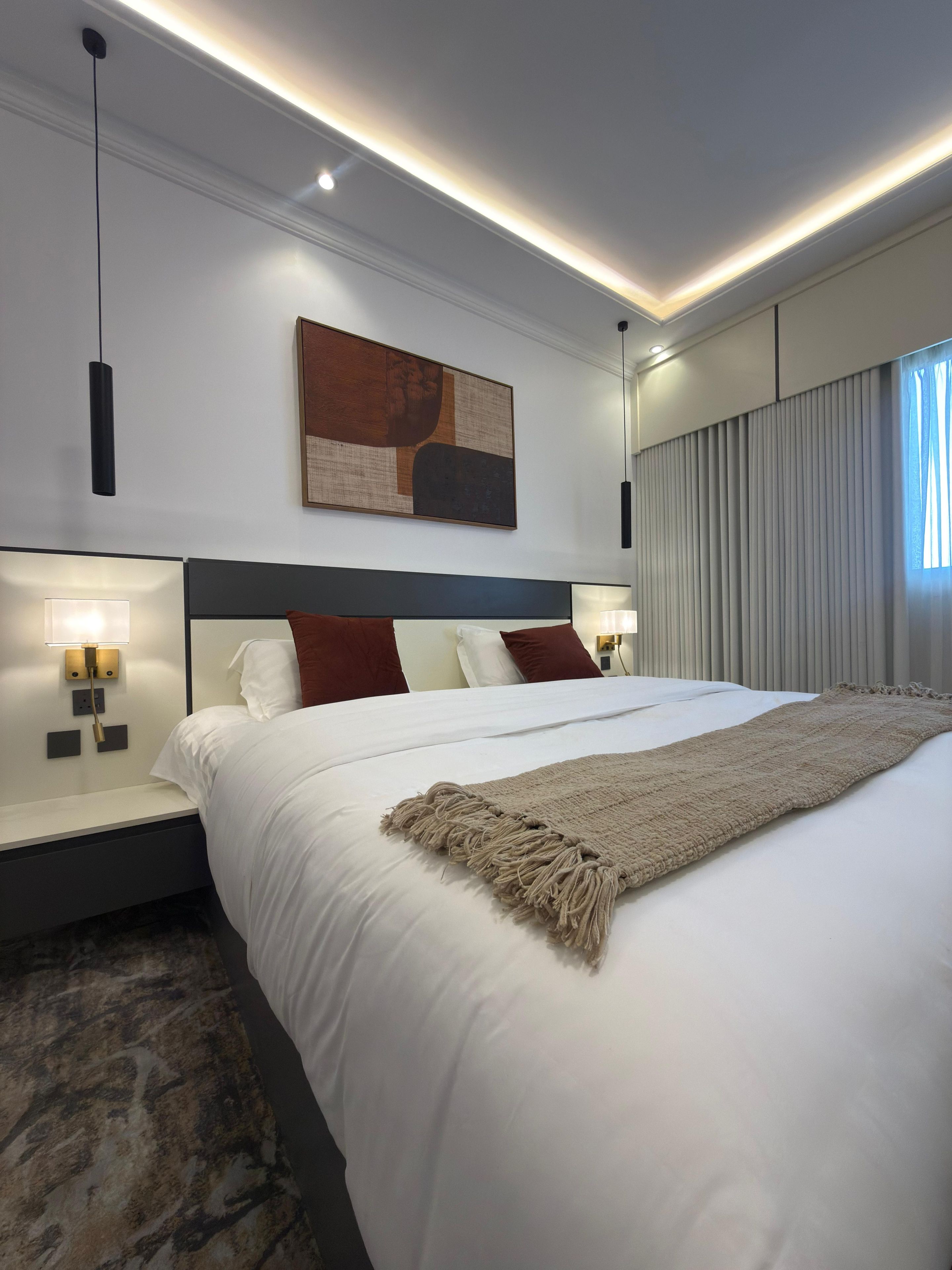 A modern hotel room with a large bed, a headboard, bedside lamps, a wall-mounted abstract painting, and a window with curtains.