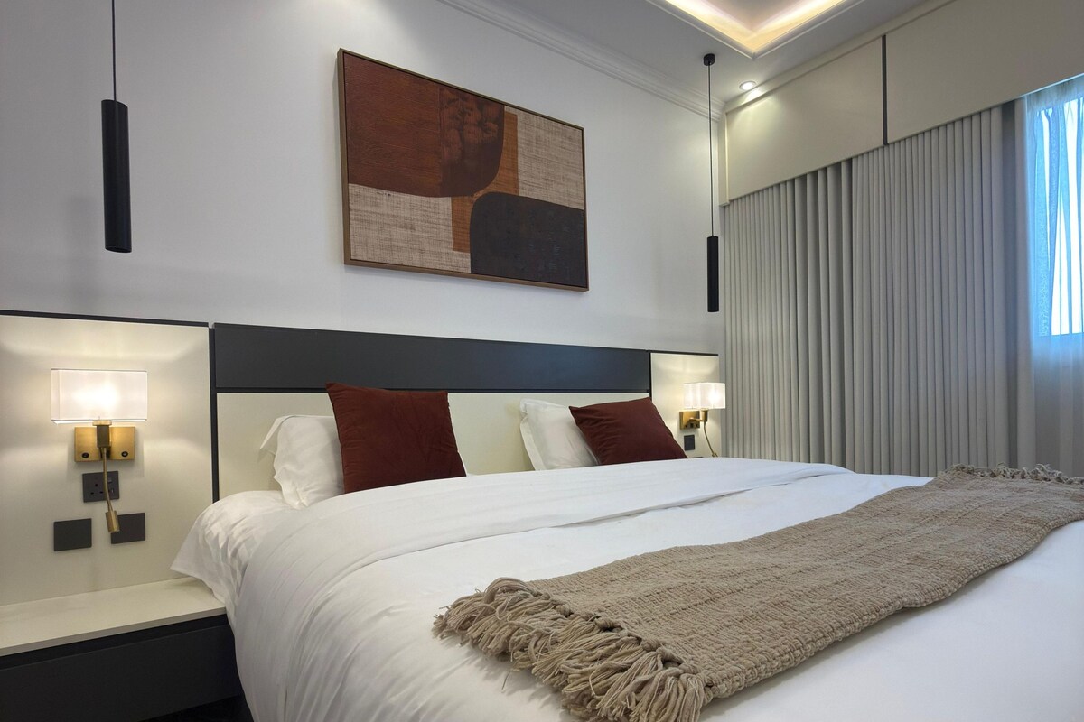 Quarto de hotel moderno com uma cama grande, cabeceira, luminárias de cabeceira, pintura abstrata na parede e uma janela com cortinas.
