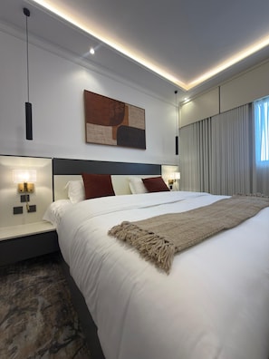 A modern hotel room with a large bed, a headboard, bedside lamps, a wall-mounted abstract painting, and a window with curtains. - Aurion Quba (Madinah)