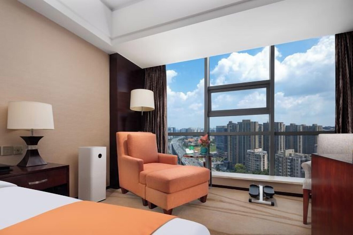 A hotel room with a bed, a chair, a desk, a lamp, and a large window with a city view.