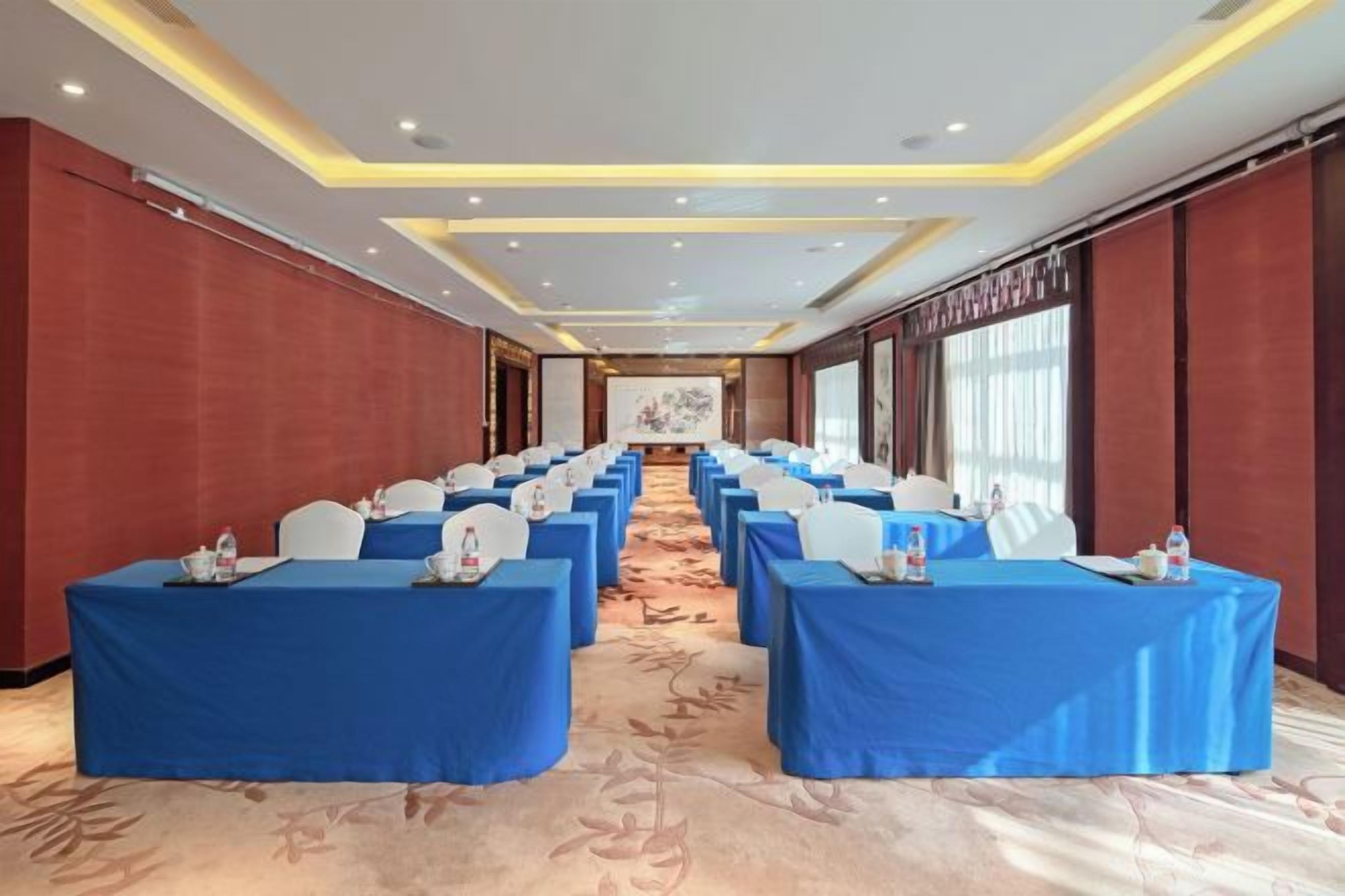 Un salón de conferencias con filas de mesas, cada una cubierta con un mantel azul y sillas blancas, y una pantalla grande al frente.