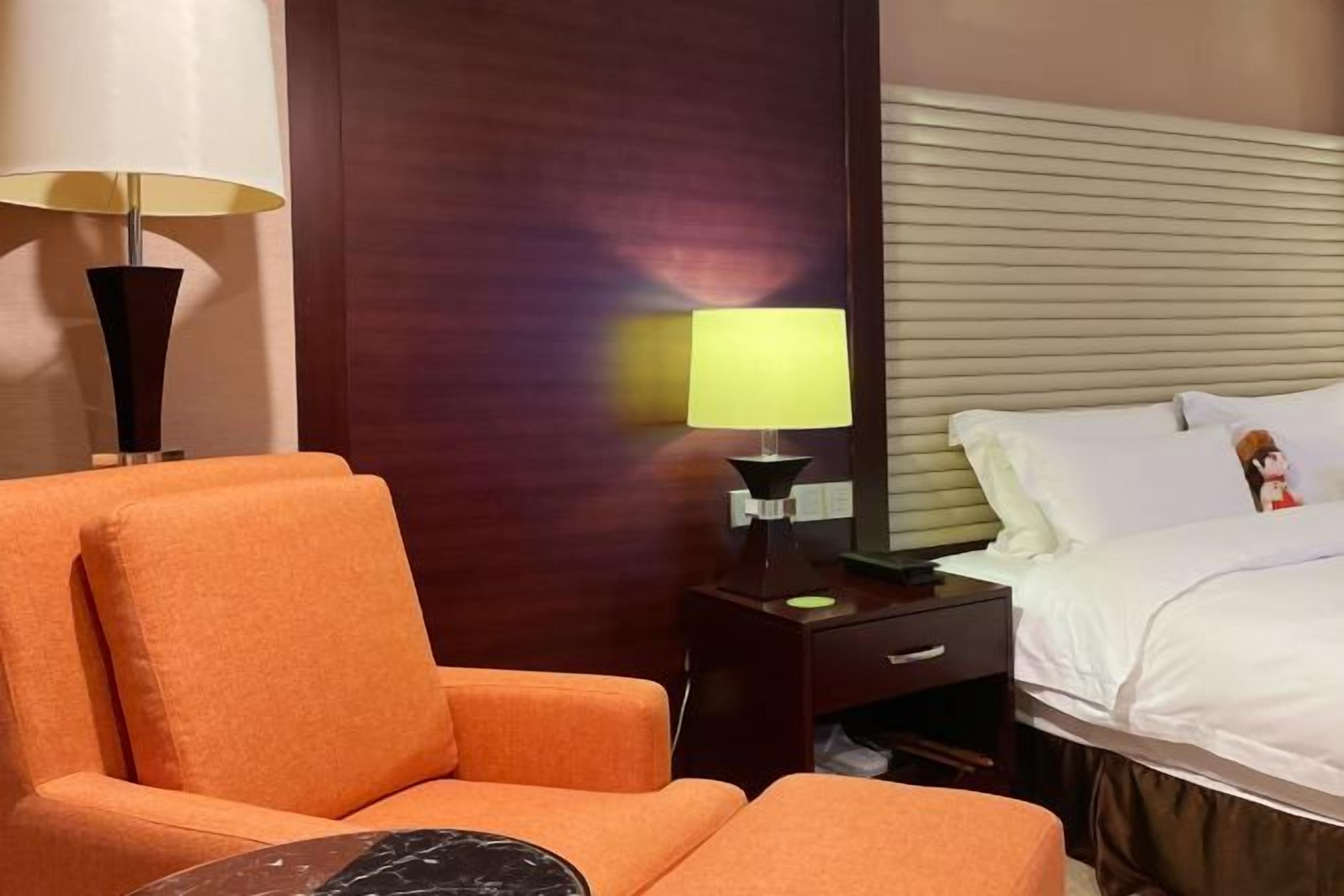 A hotel room with a bed, a nightstand with a lamp, an orange armchair, and a wooden headboard.
