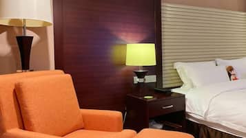A hotel room with a bed, a nightstand with a lamp, an orange armchair, and a wooden headboard.
