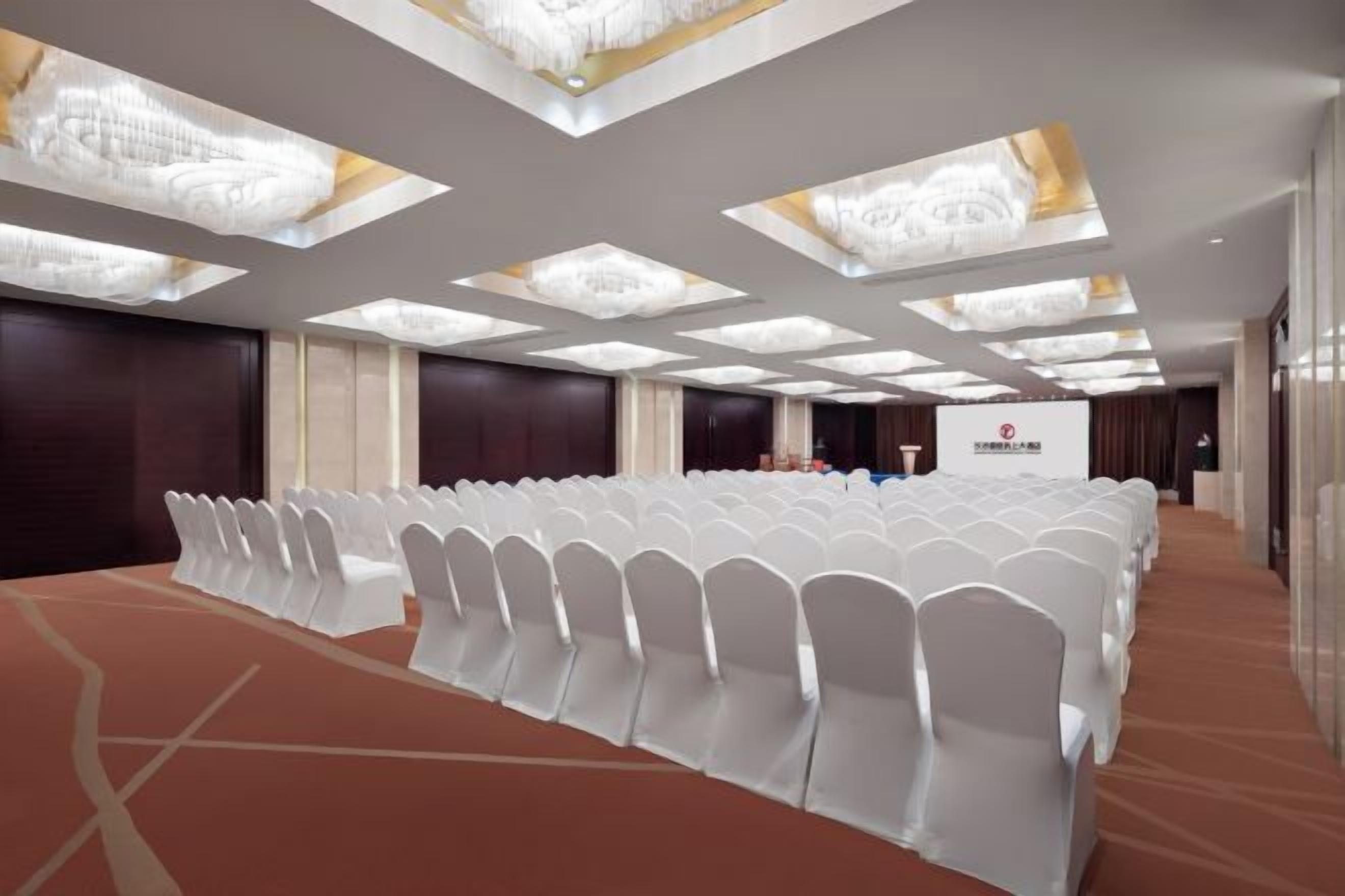 A conference room with rows of white chairs, a carpeted floor, and a ceiling with recessed lighting.