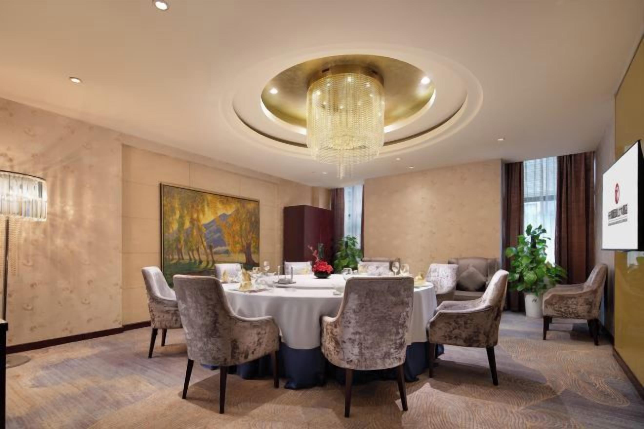 A hotel dining room with a round table, chairs, a chandelier, artwork, and a plant.