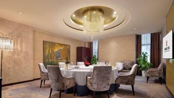 A hotel dining room with a round table, chairs, a chandelier, artwork, and a plant.
