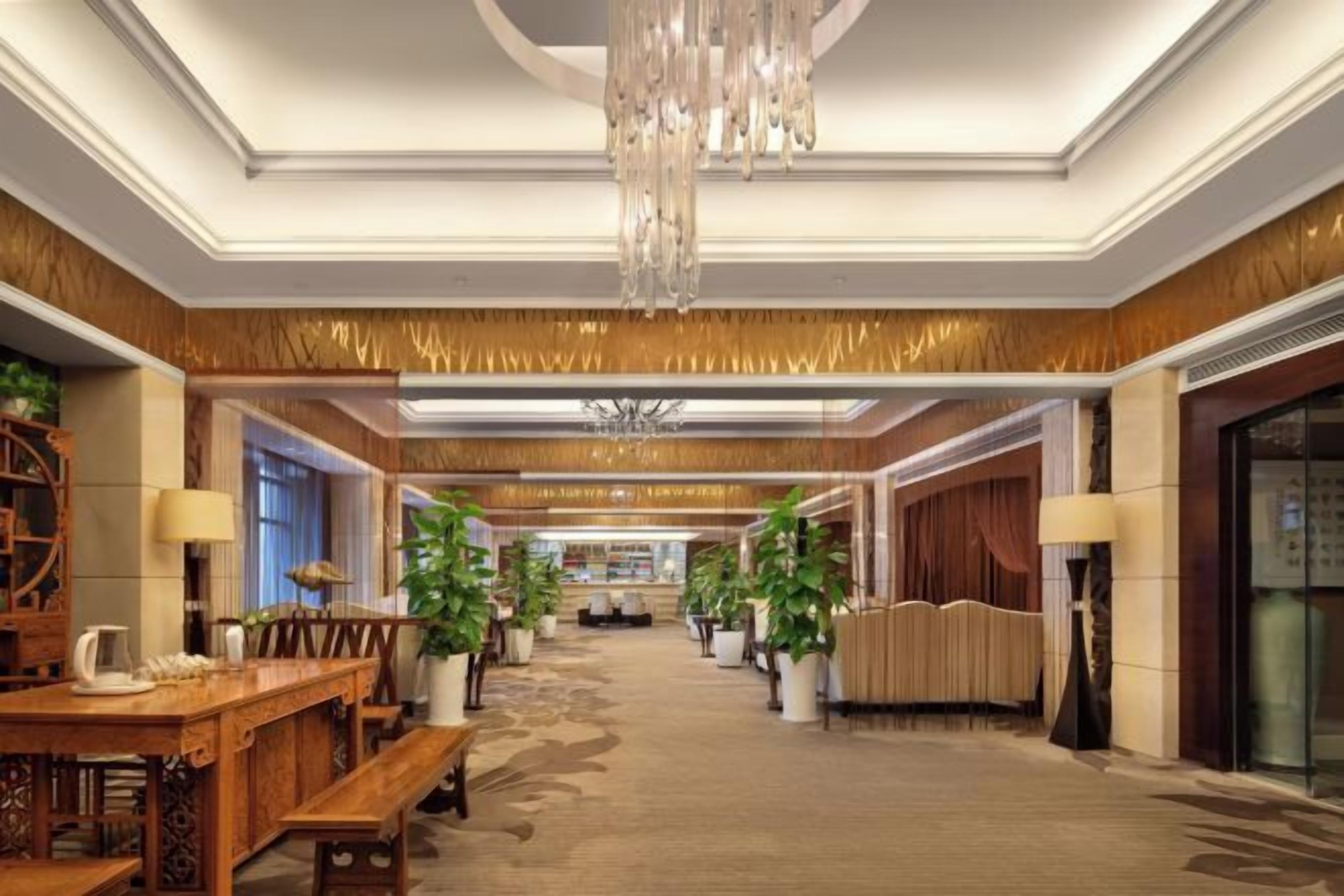 A hotel lobby with a chandelier, wooden furniture, potted plants, and a carpeted floor.