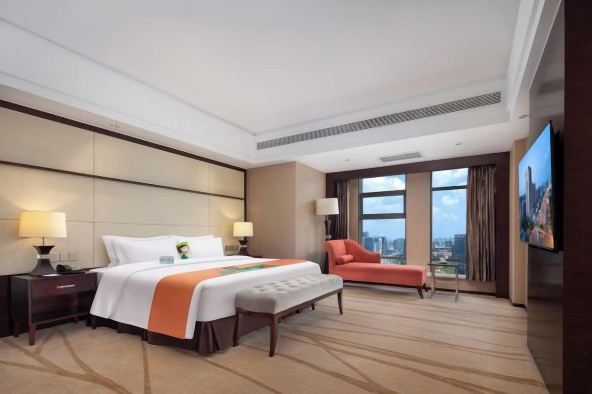 A hotel room with a large bed, a flat-screen TV, a city view, and a seating area with a chair.