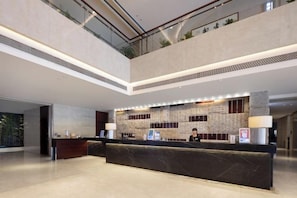 Uma área de recepção de hotel moderna com teto alto, balcão de atendimento e vista para o andar superior.