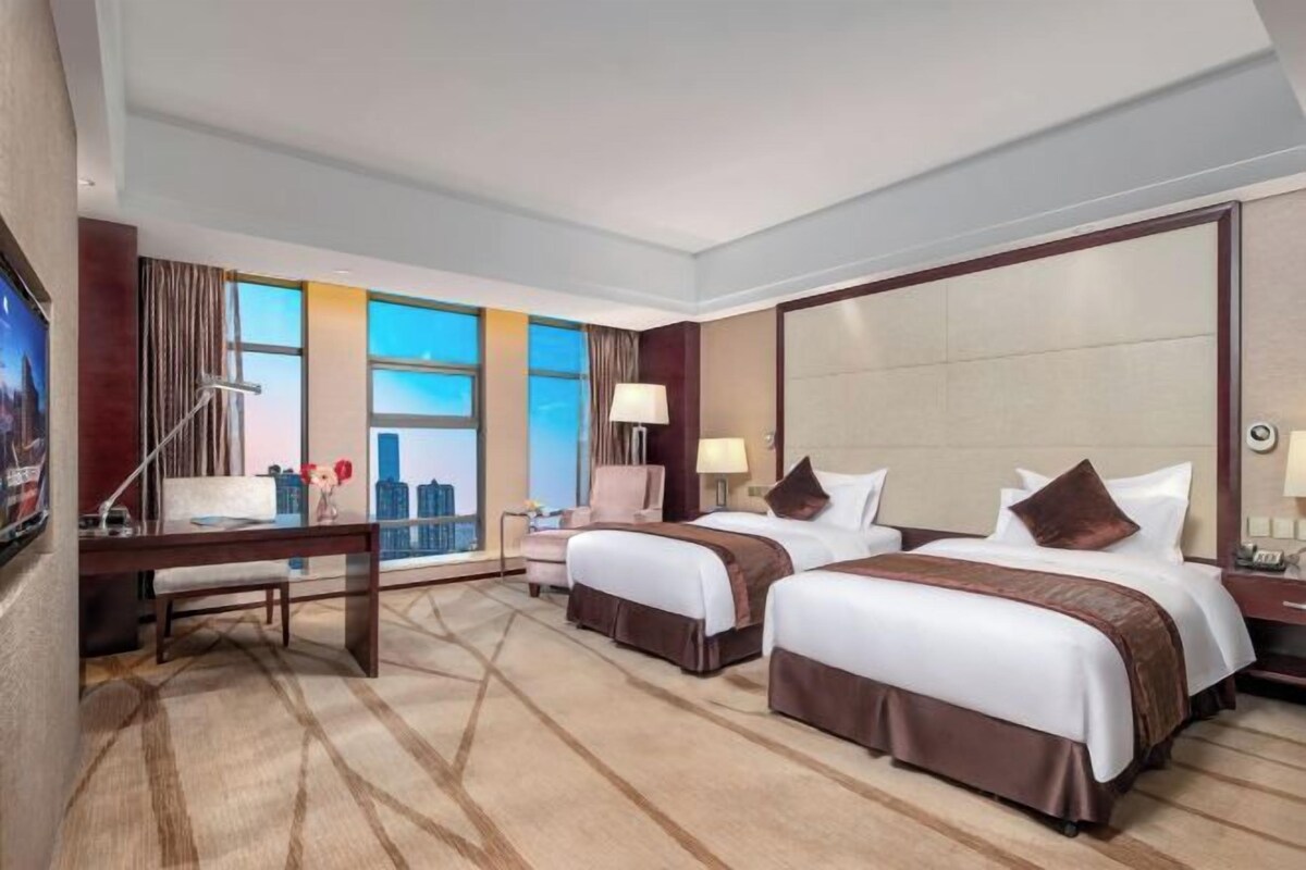 A hotel room with two beds, a desk, a chair, a TV, and a large window with a city view.
