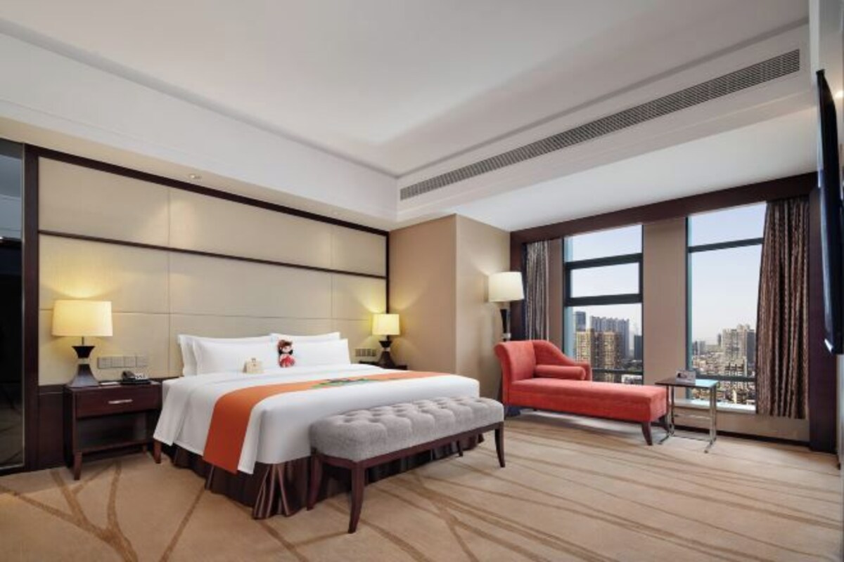 A hotel room with a large bed, a red armchair, a small table, and a city view.