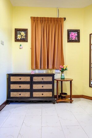 A room with a large window covered by a brown curtain, a black and beige cabinet with drawers, a small wooden table with a vase of flowers, and two framed pictures on the wall. - Classic 5BR Villa with Pool&Gardens(SP1) (Pattaya City,South Pattaya)