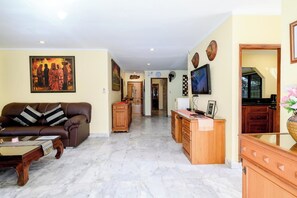 A living room with a sofa, a coffee table, a TV, and a kitchen area with cabinets and a countertop. - Classic 5BR Villa with Pool&Gardens(SP1) (Pattaya City,South Pattaya)