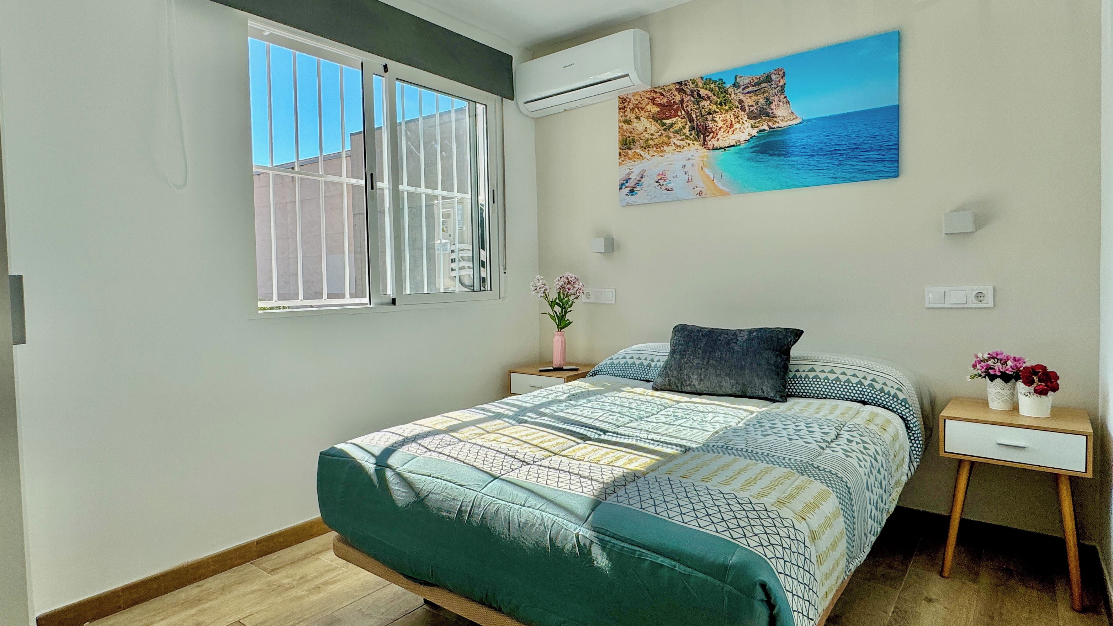 Un dormitorio con cama, mesita de noche, ventana con persianas, unidad de aire acondicionado y un cuadro con una escena de playa.