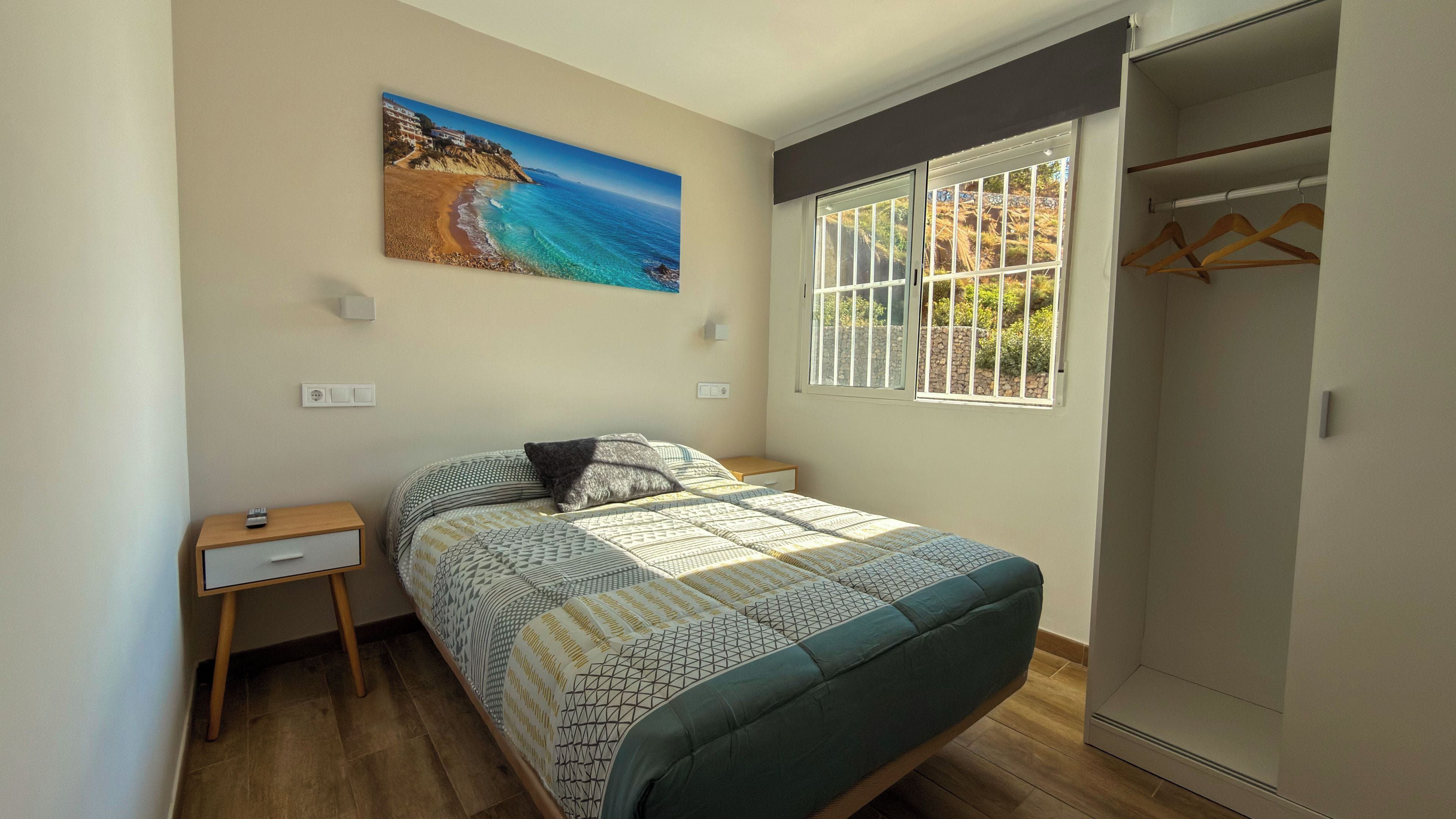 Un dormitorio con cama, mesita de noche, armario, ventana con barrotes y un cuadro con una escena de playa.