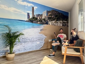 A room with a beach mural, a potted plant, and a wooden chair. - ALC HOMES SELF CHECK IN (Alacant)